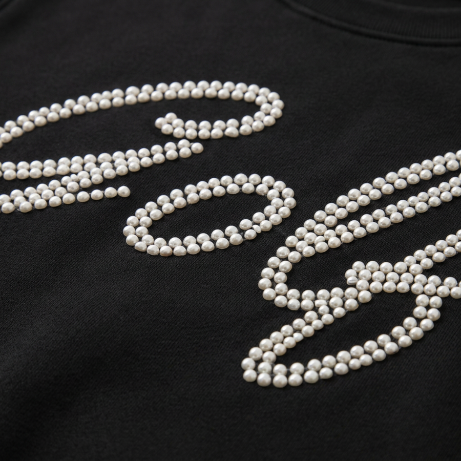 Close-up of lustrous white pearl studs forming 'Golf' script on black pullover — handcrafted pearl embellishment detail"